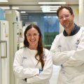 Two scientists wearing white lab coats, and goggles stand in a corridor smiling at the camera.